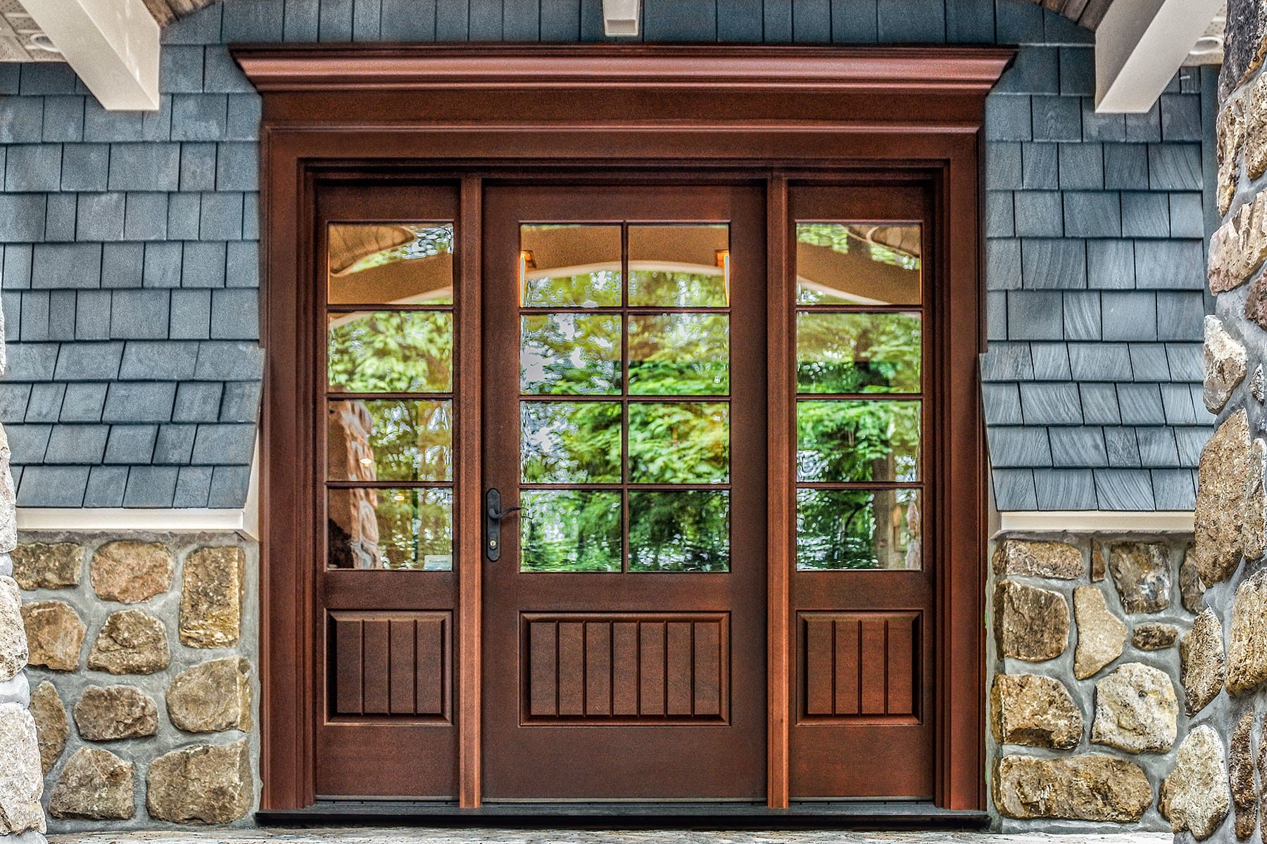 Porte d’entrée en bois Lepage Millwork au fini brun-rouge, deux latéraux vitrés à carreaux et couronnement mouluré, façade de pierre et de bardeaux