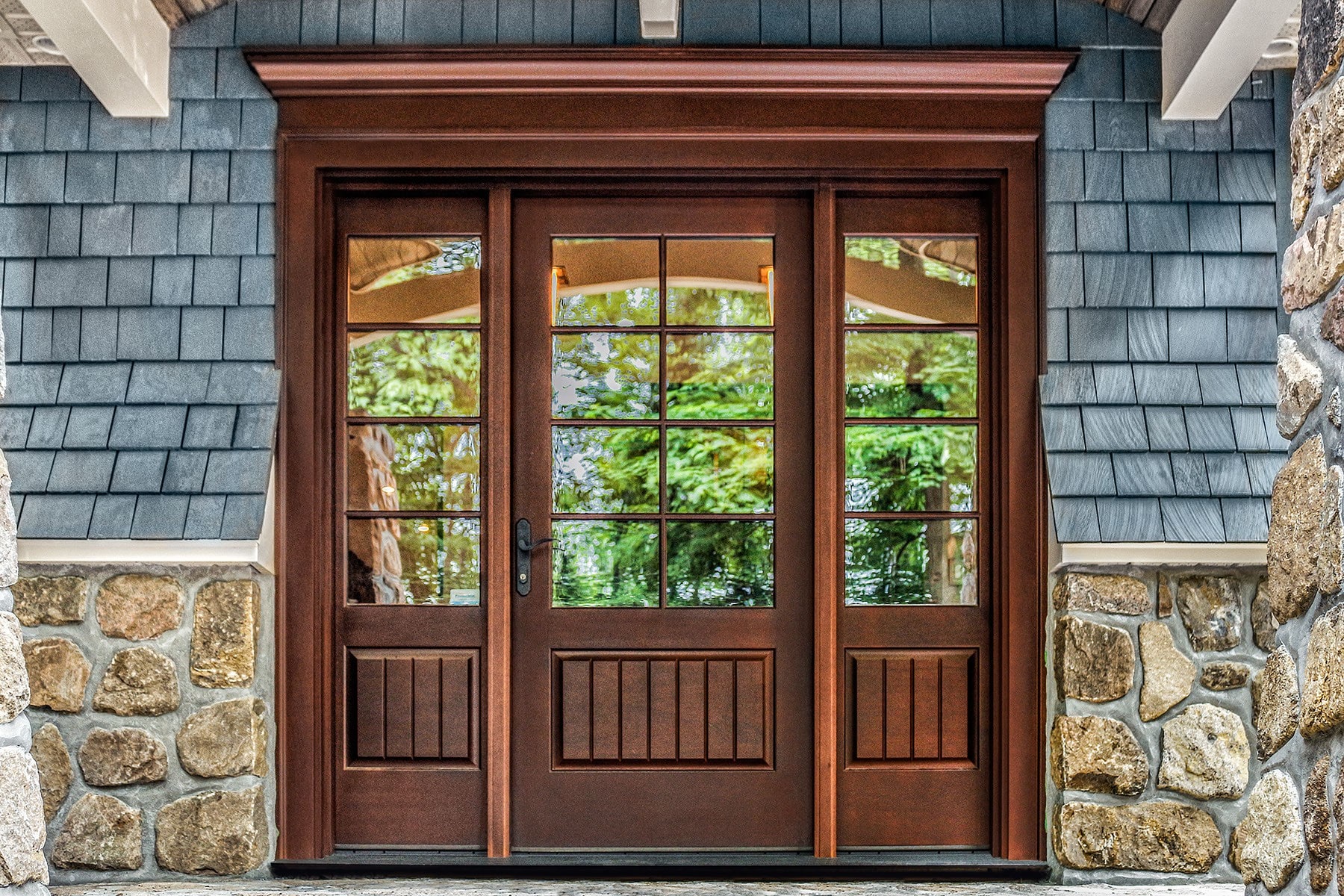 Porte d’entrée en bois Lepage Millwork au fini brun-rouge, deux latéraux vitrés à carreaux et couronnement mouluré, façade de pierre et de bardeaux